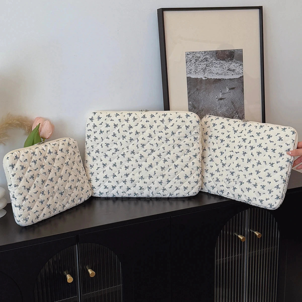 Trois pochettes matelassées à petites fleurs bleues, en trois tailles, alignées sur un buffet noir devant un cadre photo.