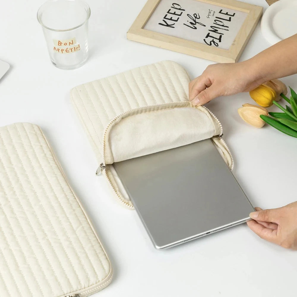 Pochette matelassée en tissu écru pour ordinateur portable, ouverte, laptop gris posé dessus dans une mise en scène de bureau lumineuse avec tulipes jaunes.