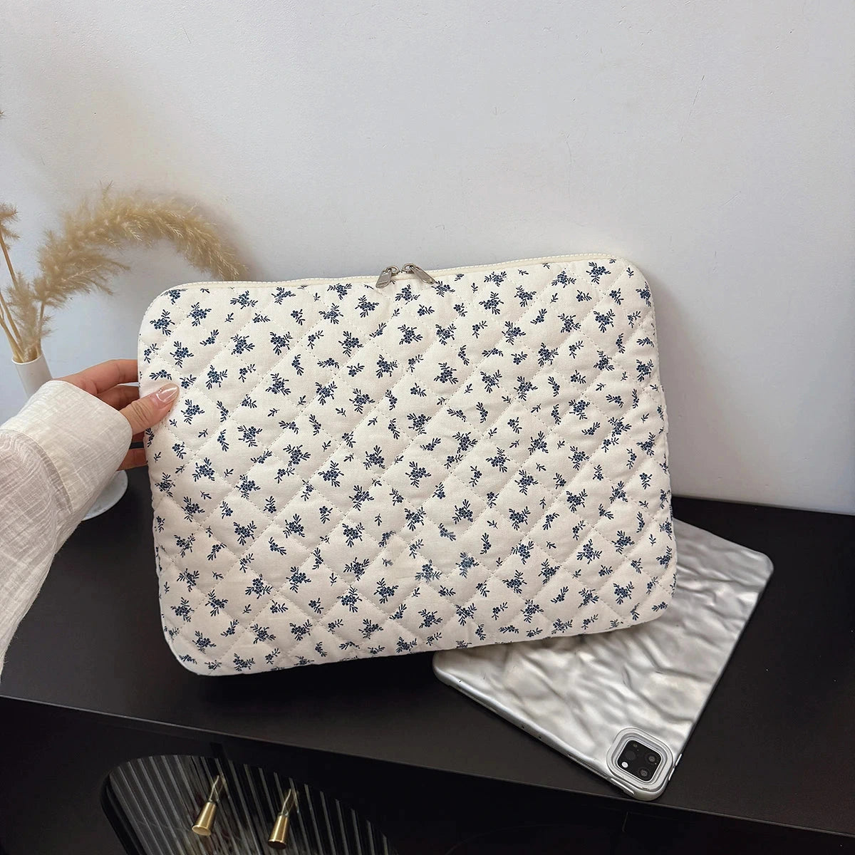Pochette matelassée en tissu écru à petites fleurs bleues, tenue à la main devant une tablette posée sur un bureau noir.