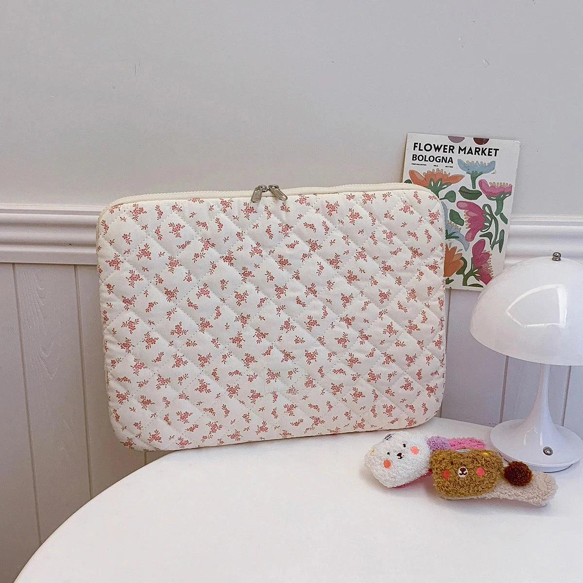 Pochette ordinateur matelassée blanc cassé à motifs floraux roses, fermeture éclair, tirette dorée, posée sur une table.