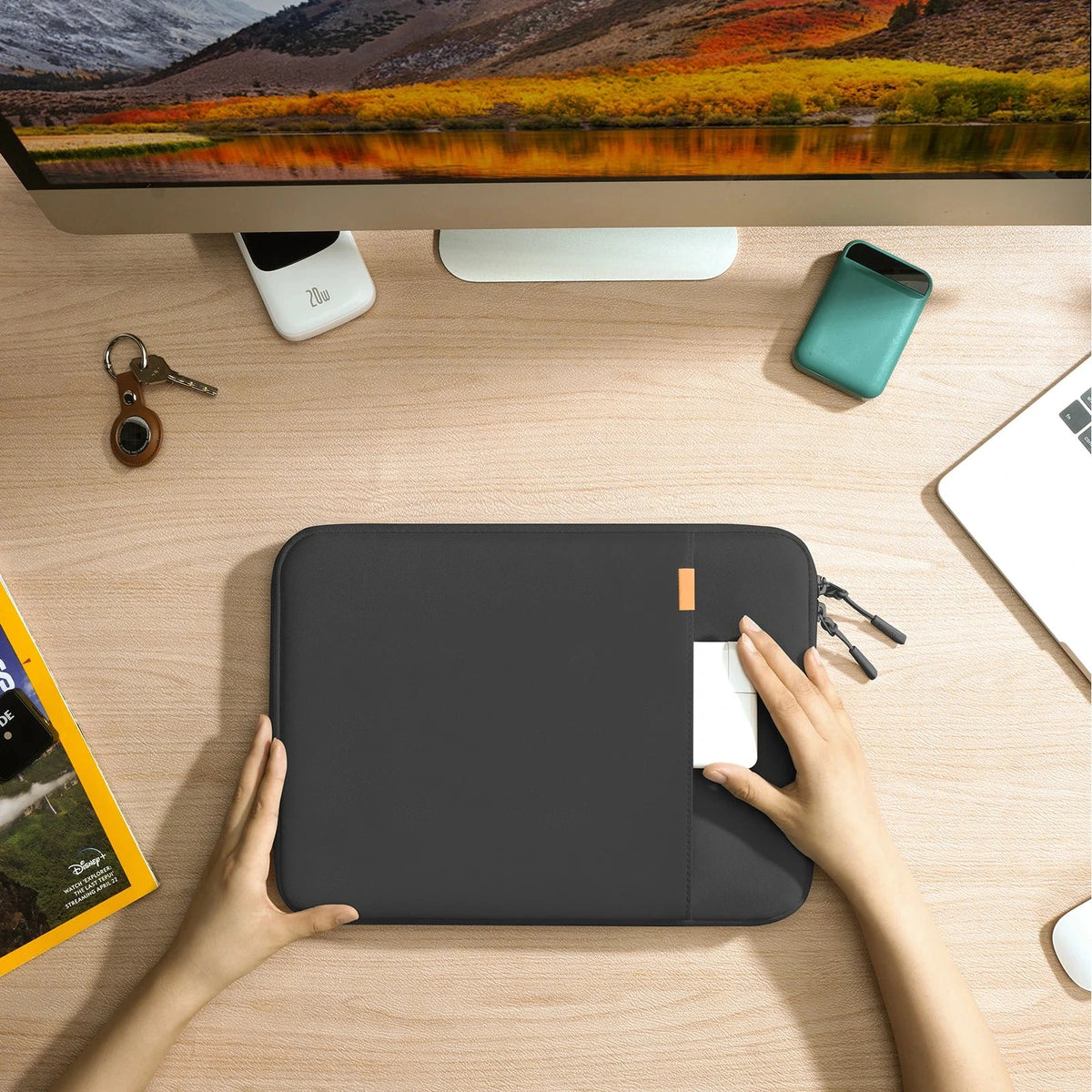 Vue de dessus d'un bureau : deux mains glissent un ordinateur portable blanc dans la pochette noire posée sur le plan de travail.