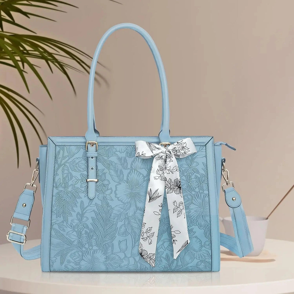 Sac à main bleu clair en simili cuir gaufré de motifs floraux, deux anses, foulard blanc à fleurs noué sur l'anse droite, bandoulière amovible, posé sur un plateau clair.