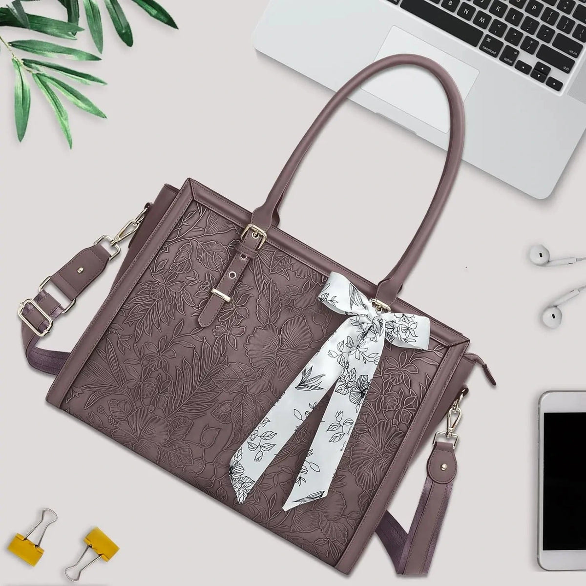 Grand sac ordinateur prune en similicuir gaufré de fleurs, foulard imprimé noué à l'anse, posé sur un bureau avec un ordinateur portable, des écouteurs et un téléphone.