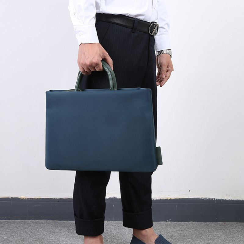 Homme en chemise blanche et pantalon sombre tenant à la main une sacoche pour ordinateur bleu pétrole à poignées vertes.