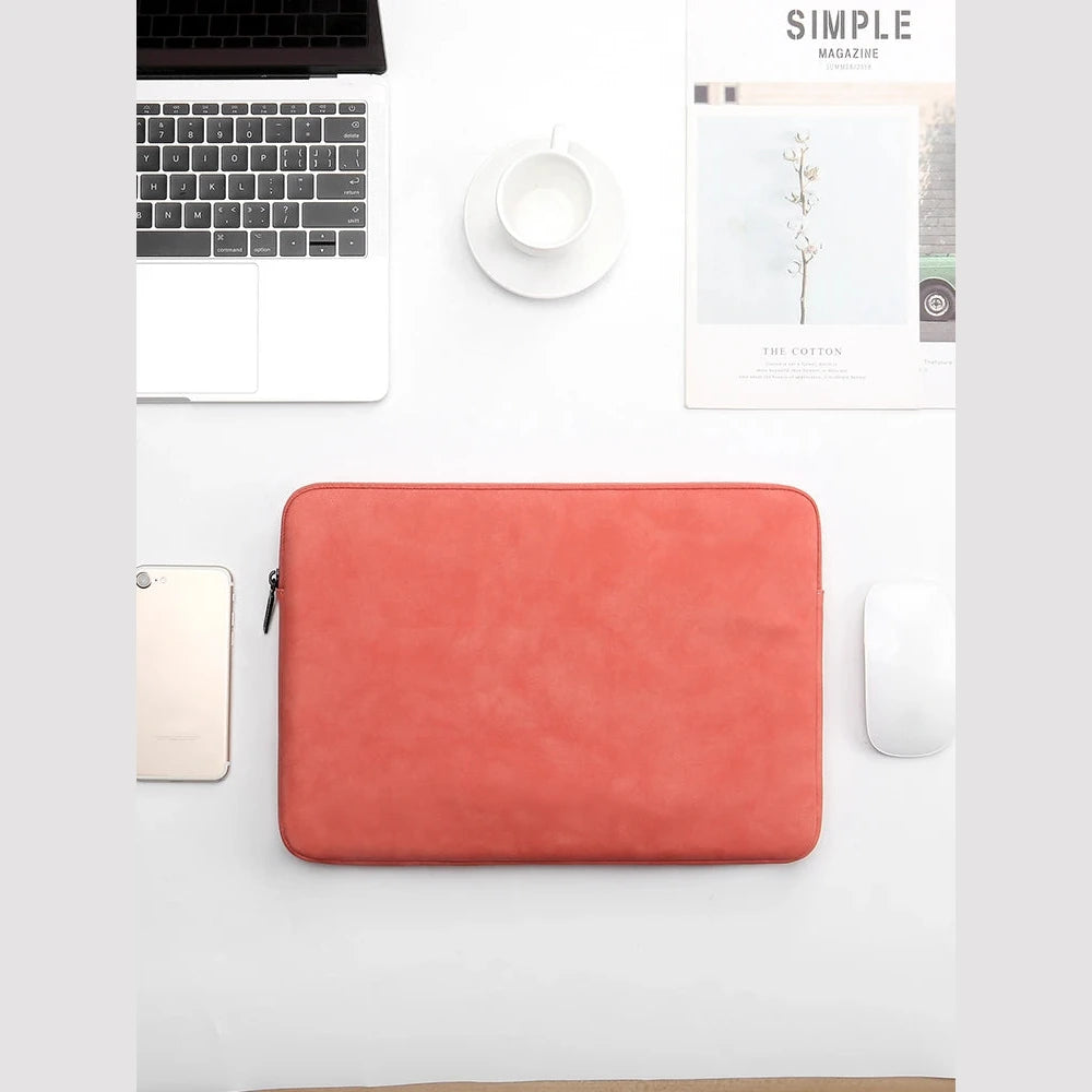 Housse rouge corail pour ordinateur portable posée sur un bureau blanc, entre un ordinateur ouvert, une tasse de café et un magazine.