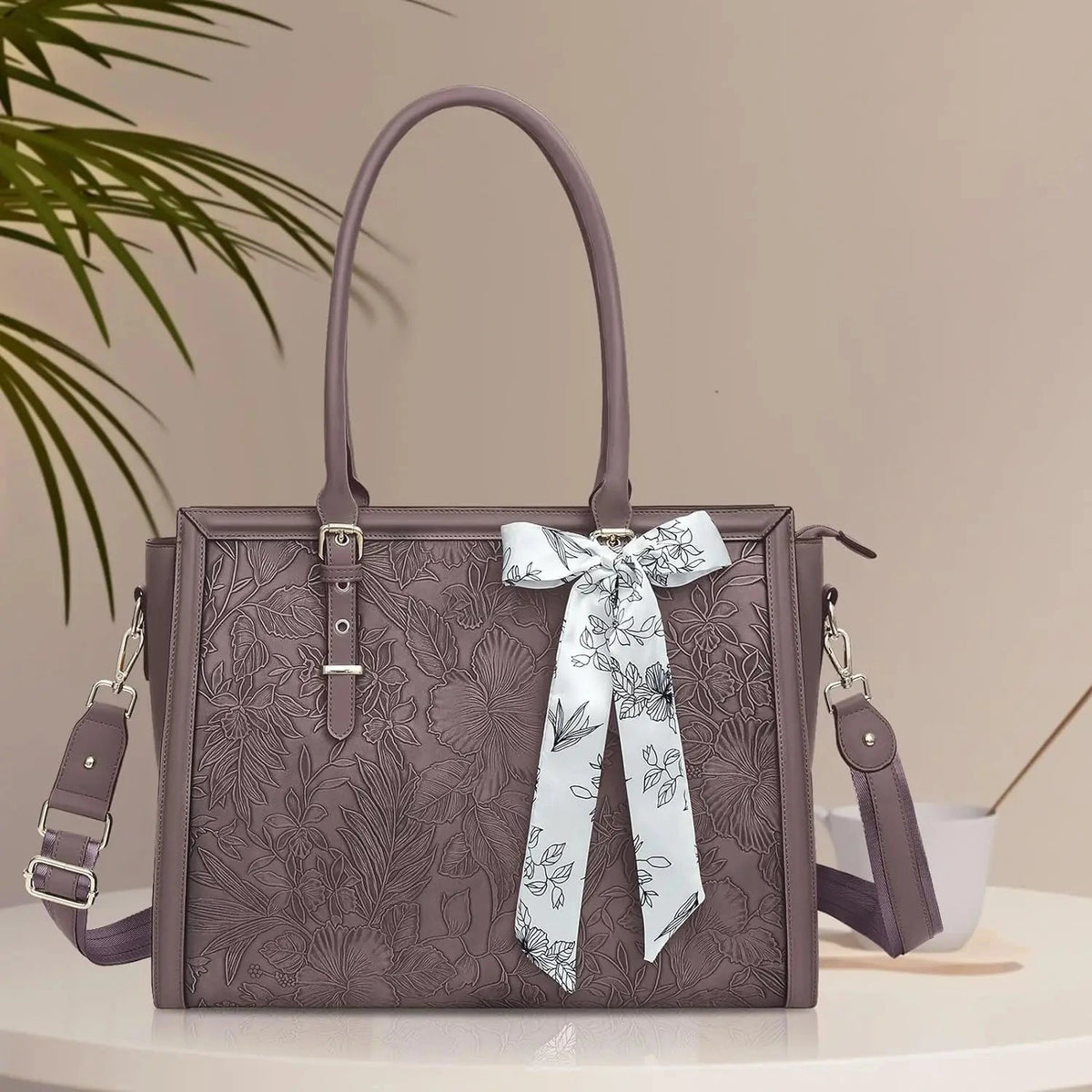 Grand sac ordinateur prune en simili cuir gaufré de motifs floraux, deux anses, bandoulière réglable amovible et foulard blanc à fleurs noué sur l'anse.