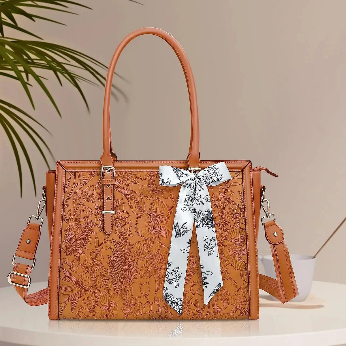 Grand sac ordinateur camel à motif floral gaufré, vu de face avec son foulard noué, posé sur un plateau clair devant un feuillage.