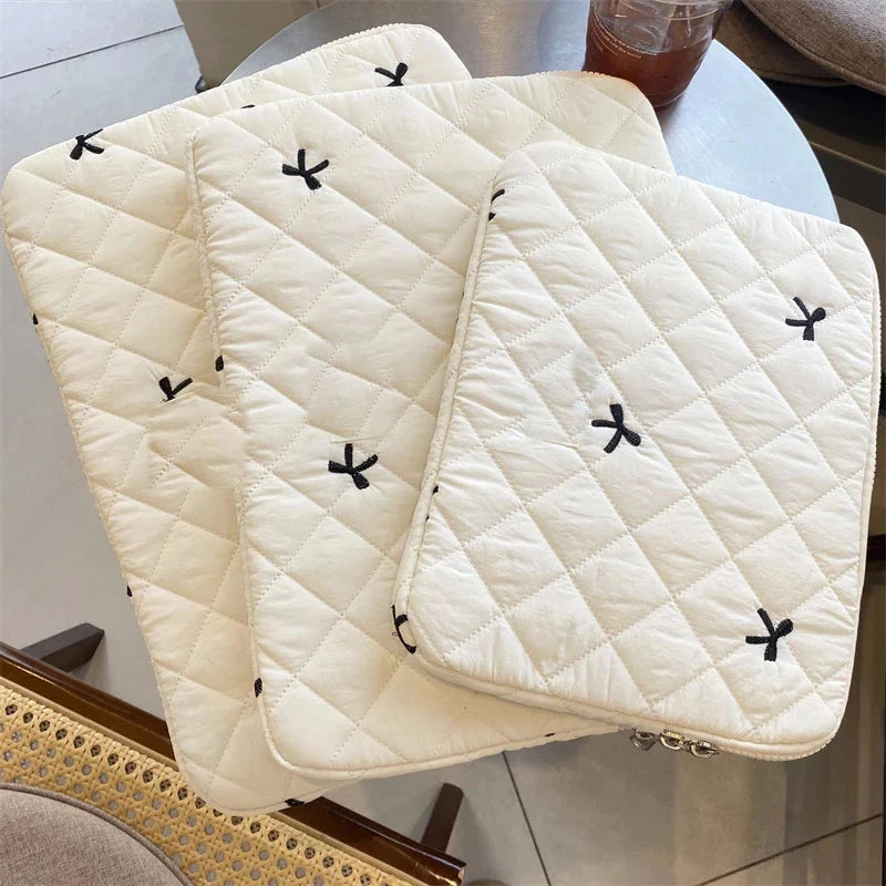 Trois housses matelassées écrues pour ordinateur portable, brodées de petits nœuds noirs, empilées sur une table ronde.