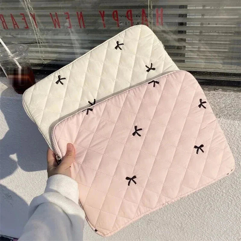 Housse matelassée rose pour ordinateur portable, brodée de petits nœuds noirs, tenue à la main devant une vitrine, une housse écrue posée derrière.